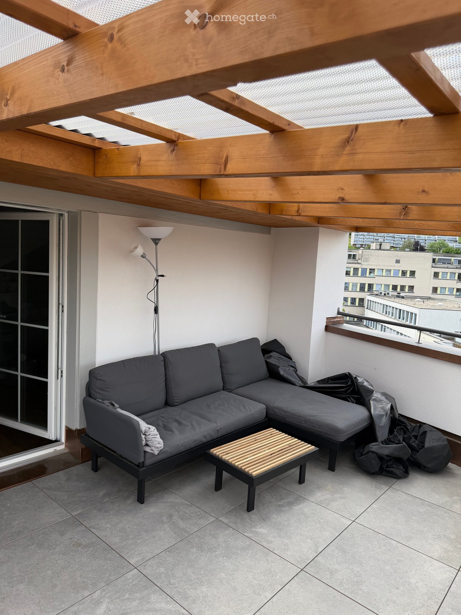 Balcony with grey sectional sofa, small wooden coffee table, floor lamp, and view of city buildings.