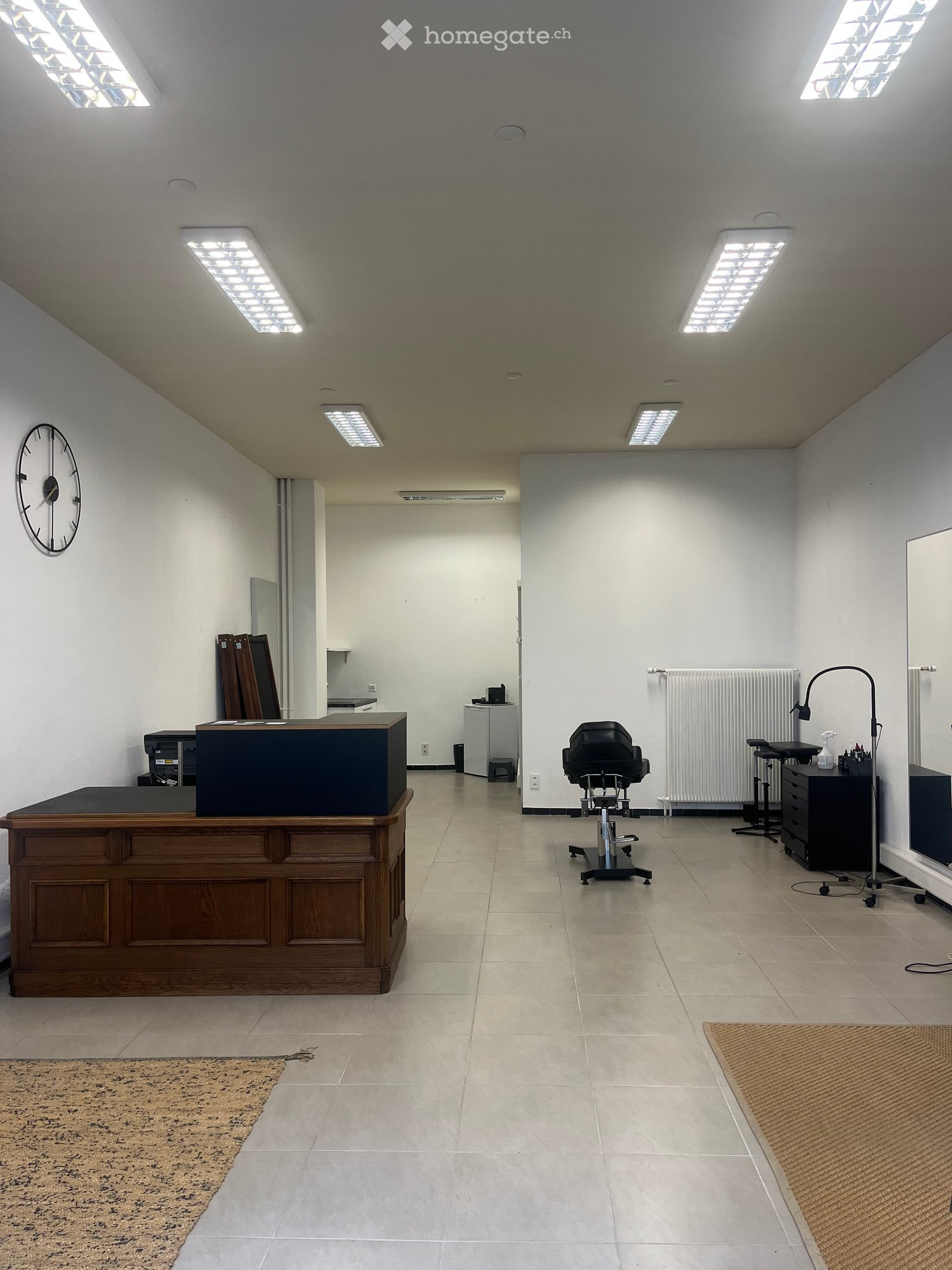 Bright, open room with a reception desk, two chairs, a mirror, a radiator, a lamp, and a clock on the wall.