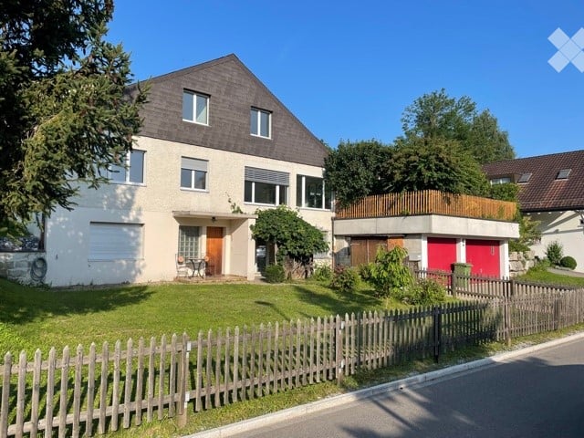 Two-story house with a modern design, featuring a dark roof, white walls, and a garage with red doors. The front yard is enclosed by a wooden fence and includes a small patio area with outdoor furniture.