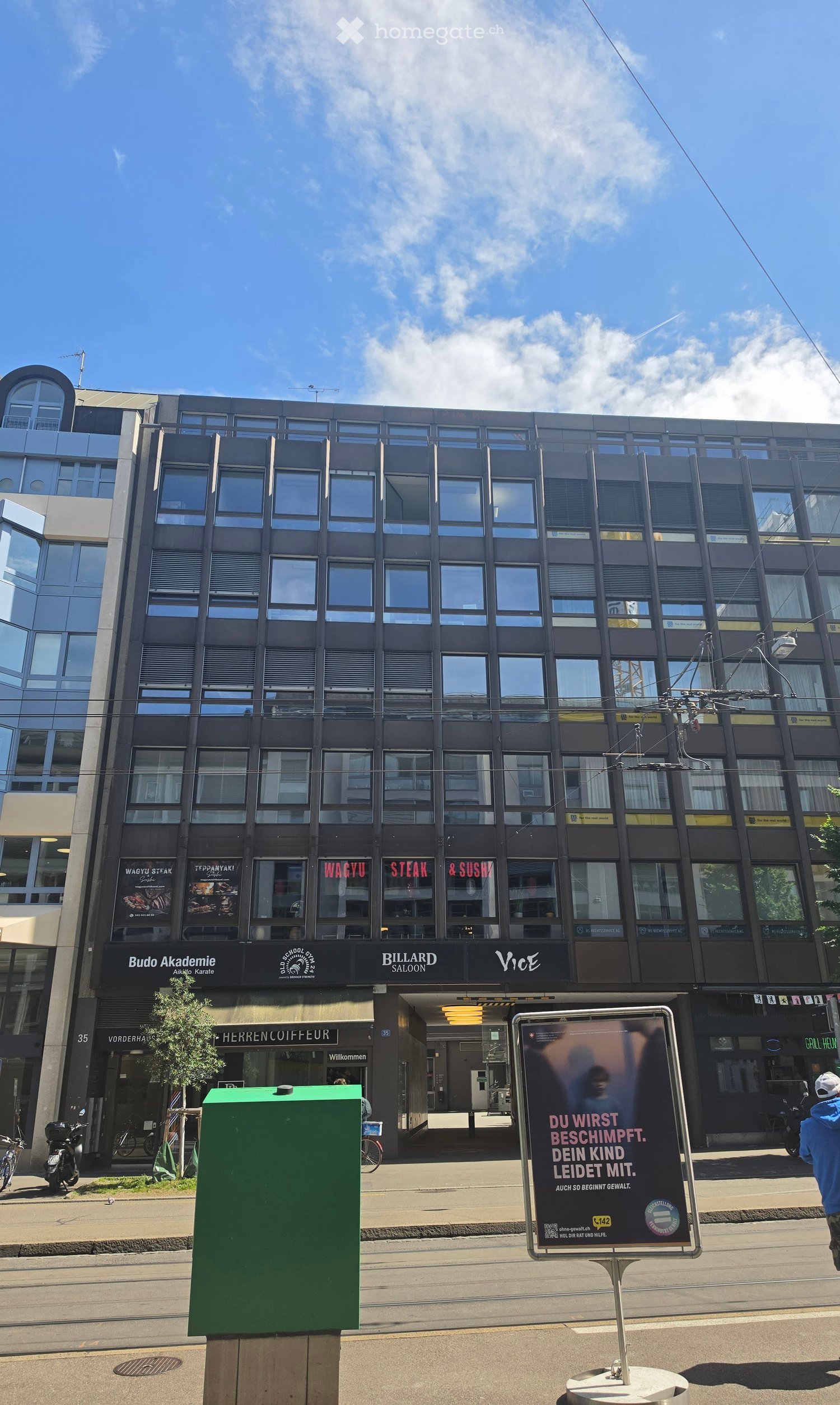Modern multi-story building with large windows and signage for various businesses including "Budo Akademie," "Billiard Saloon," and "Vice." The building has a sleek, contemporary design with reflective glass windows and a mix of commercial and possibly re