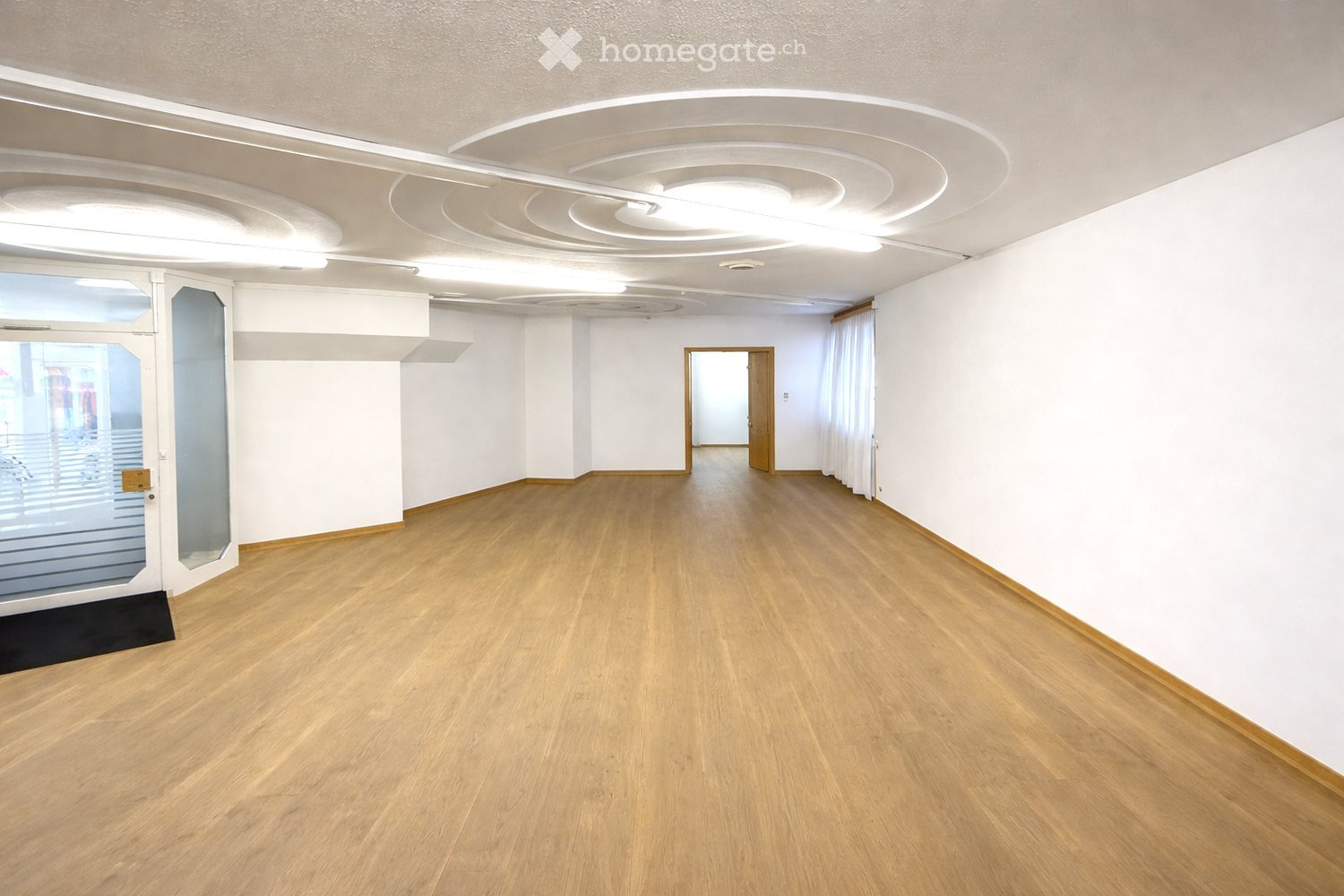 Empty room with light wooden flooring, white walls, and a glass door leading to an outdoor area. The ceiling features modern lighting fixtures.