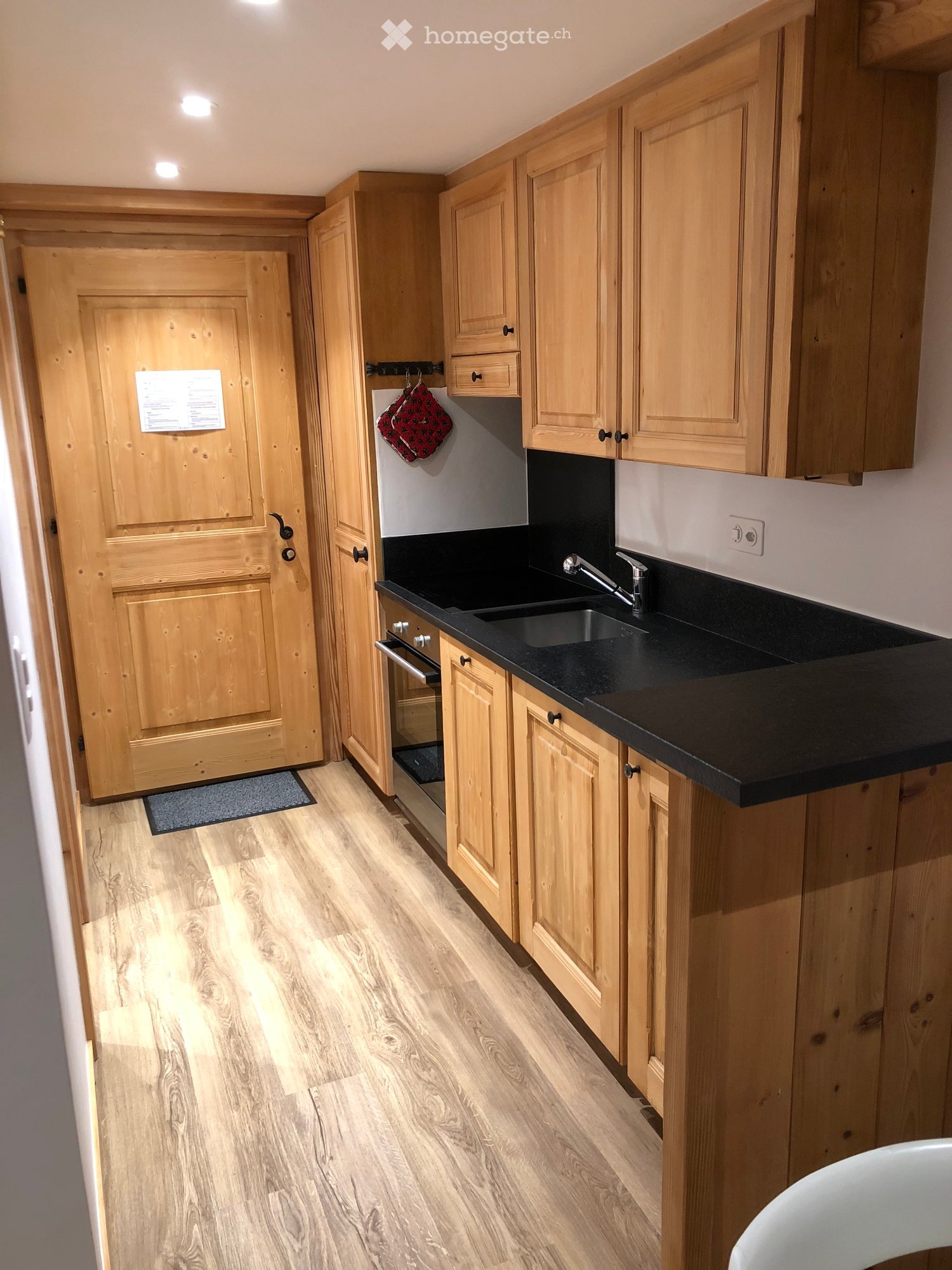 Modern kitchen with light wooden cabinets, black countertop, stainless steel sink, and faucet. Wooden floor and a small rug near the door.
