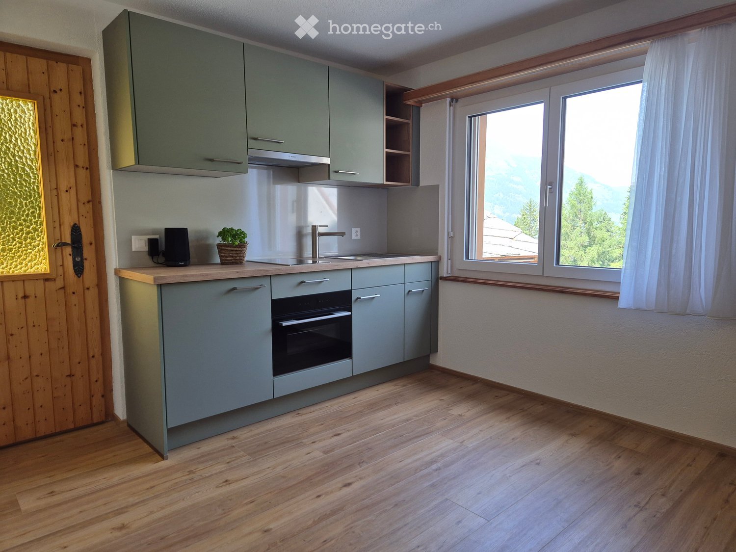 Modern kitchen with grey cabinets, wooden countertops, sink, oven, and a small plant on the counter. Large windows with white curtains offer a scenic mountain view.