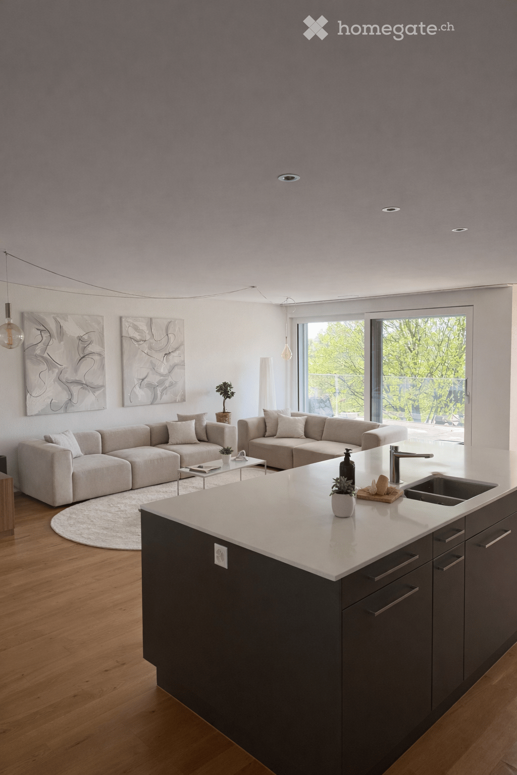 Modern open-plan living and kitchen area with a grey sectional sofa, coffee table, kitchen island, and large windows leading to a balcony.