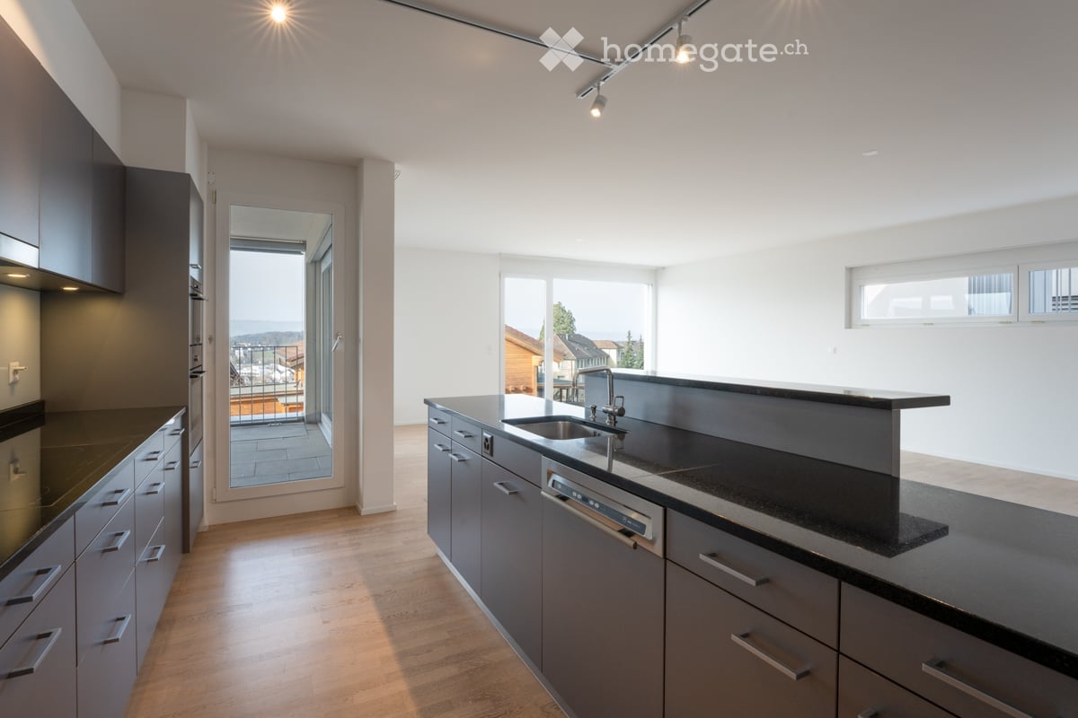 Modern kitchen with grey cabinets, black countertops, stainless steel sink, and dishwasher. Large windows and sliding doors lead to a balcony with a scenic view.