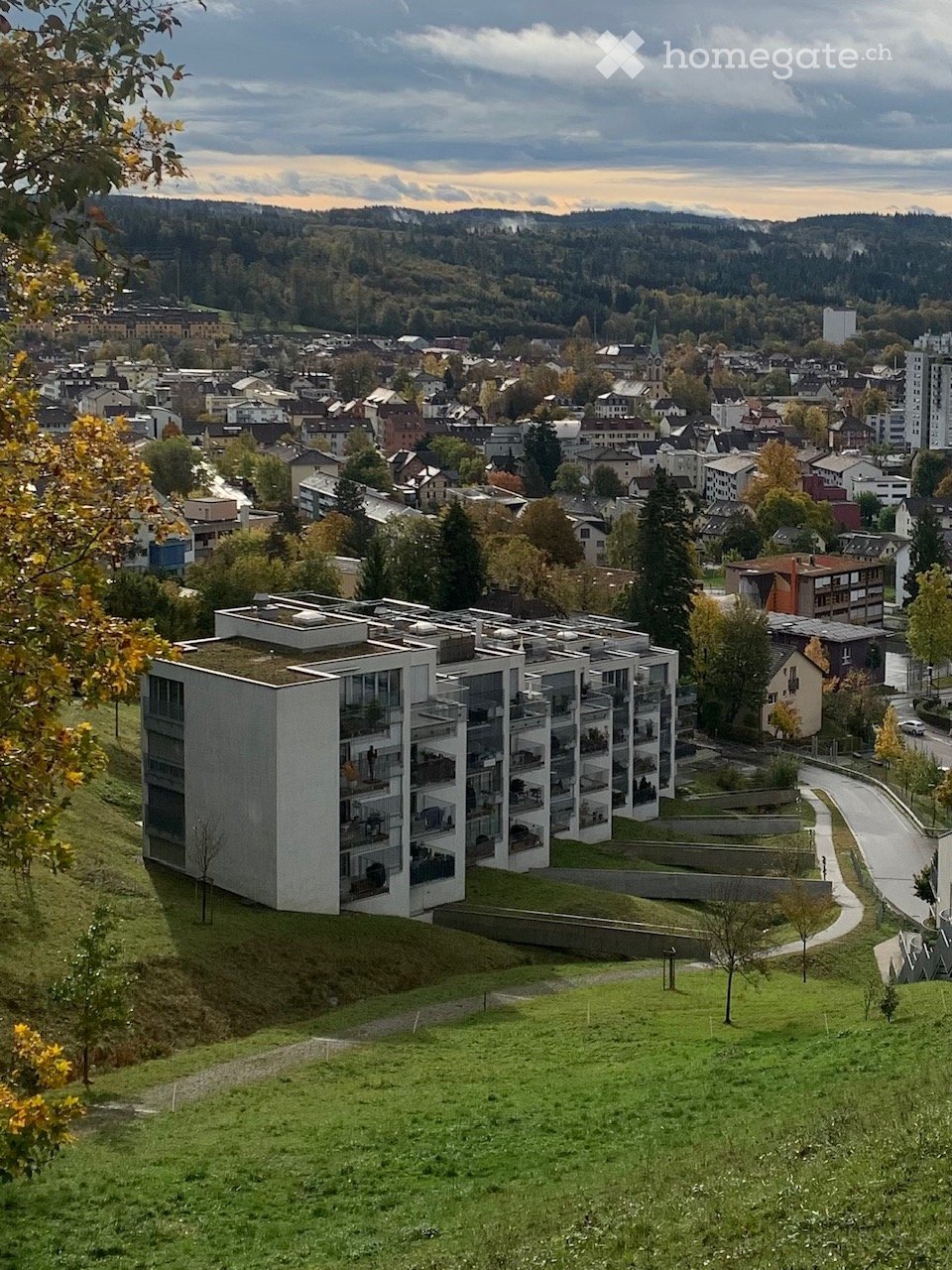 Modern multi-story residential building with balconies and green roof, situated on a hillside overlooking a town with houses, trees, and distant mountains.