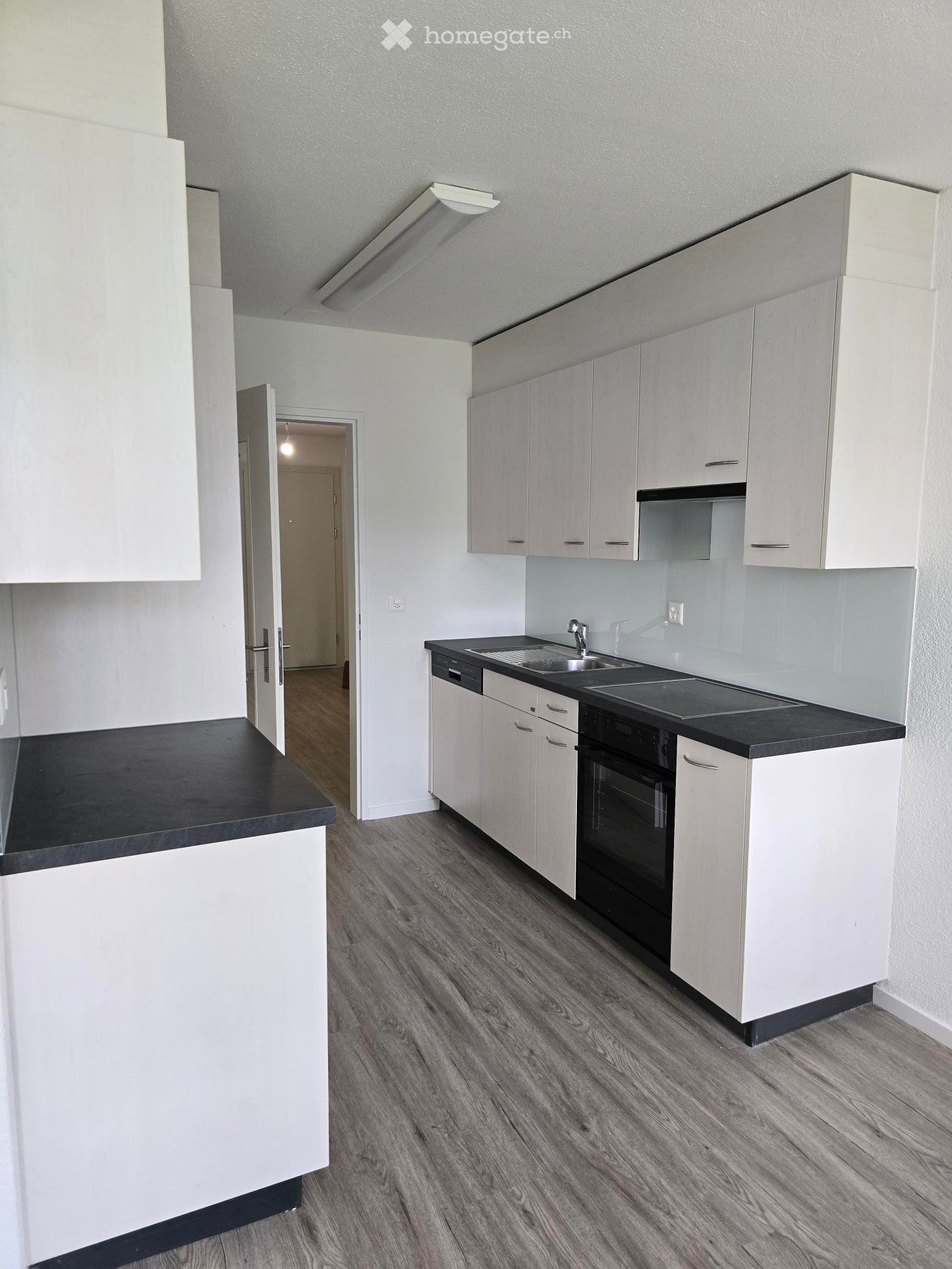 Modern kitchen with white cabinets, black countertops, and a stainless steel sink. The floor is made of wood, and there is an open doorway leading to another room.