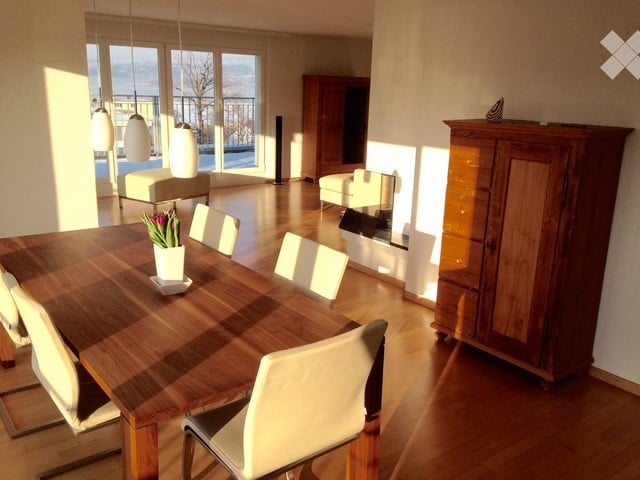 Bright, modern dining area with a large wooden dining table, white chairs, a cabinet, and a view of the balcony. The room features a fireplace and a sofa in the background.