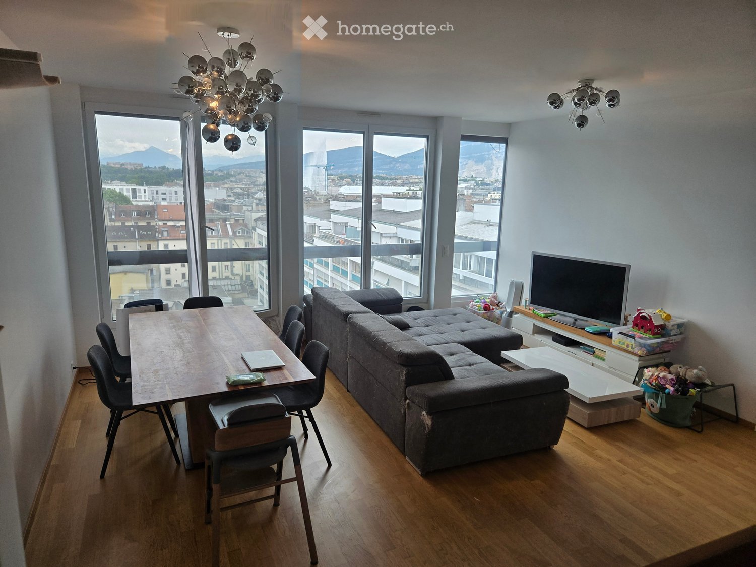 Modern living room with large windows offering a cityscape view, featuring a grey sectional sofa, a wooden dining table with chairs, a TV stand with a flat-screen TV, and decorative items.