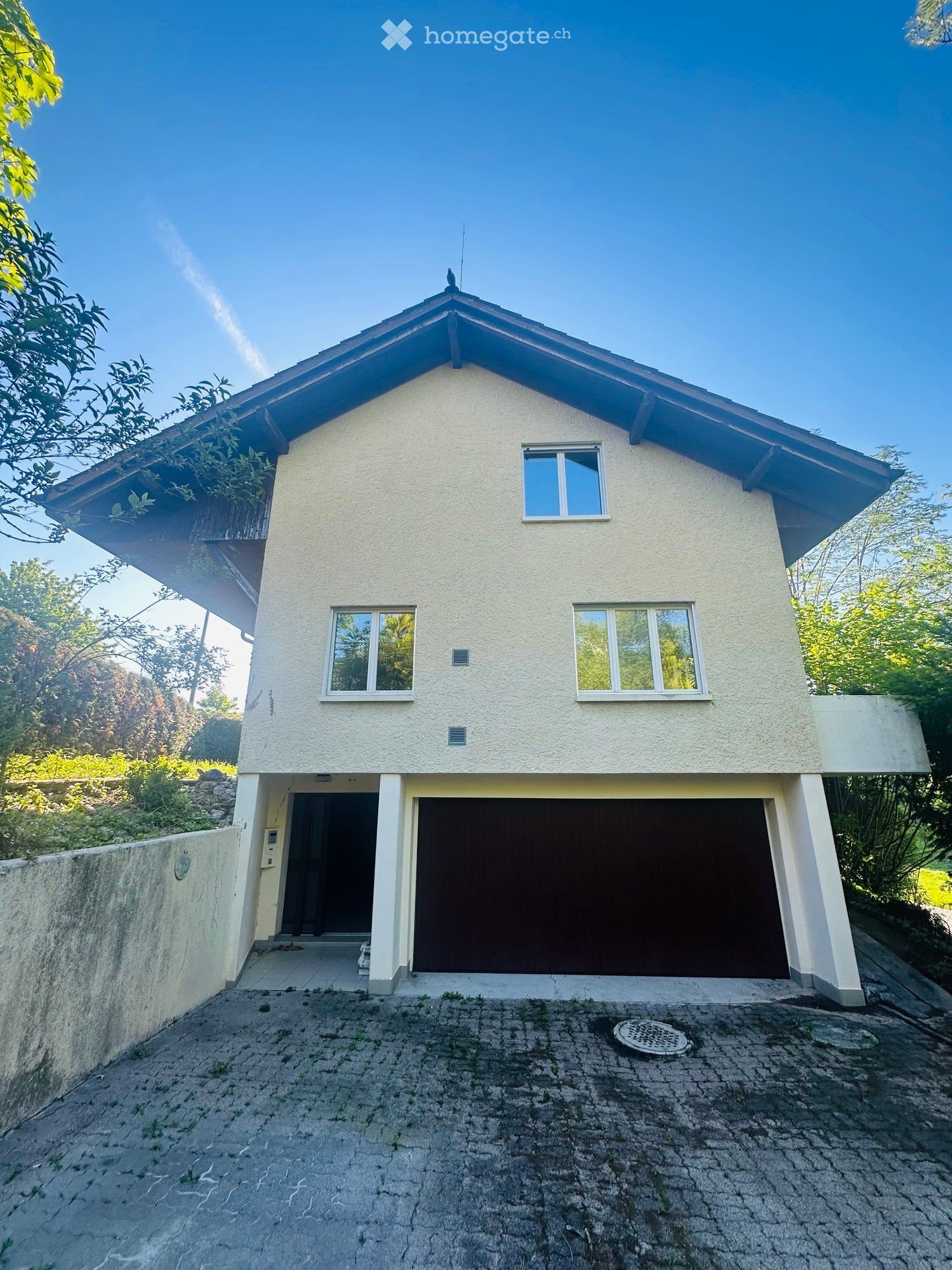 Two-story house with a raised ground floor, garage, and modern design. The house features a light-colored exterior, multiple windows, and a sloped roof.