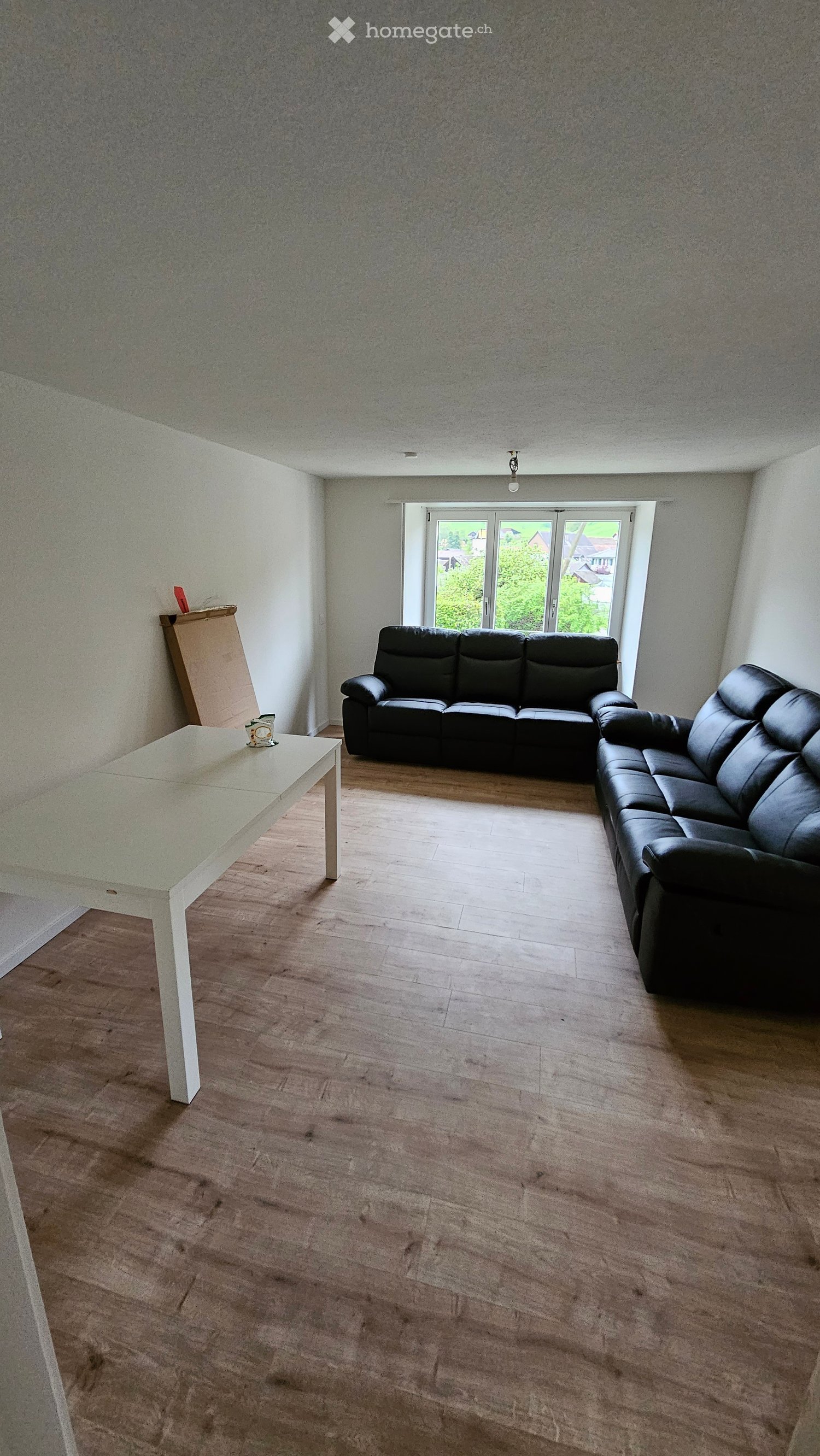 Living room with wooden flooring, white walls, and large windows. Features a black leather sofa and a white table.