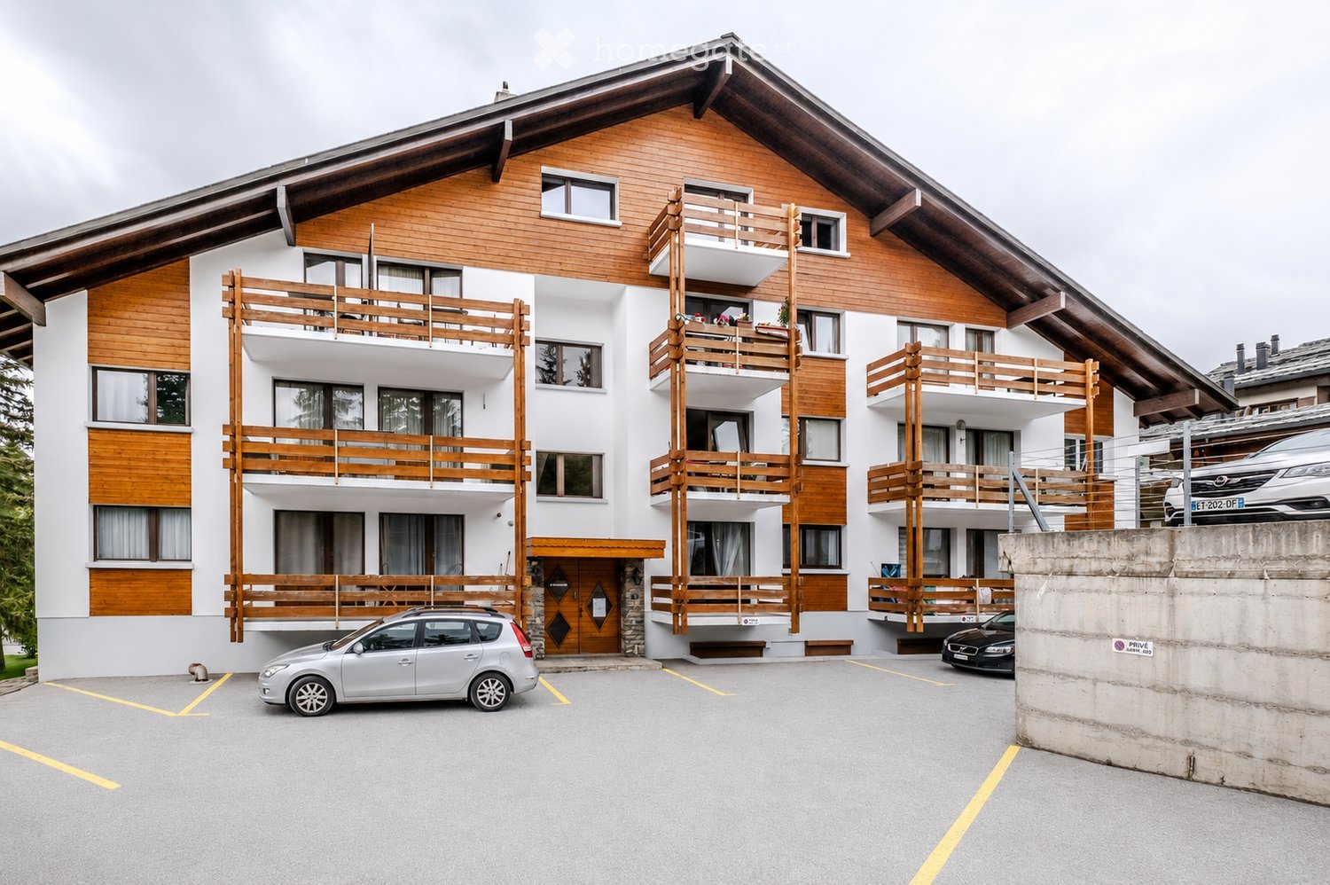A multi-story building with a wooden facade and balconies, featuring a mix of white and wooden panels, large windows, and a sloped roof.