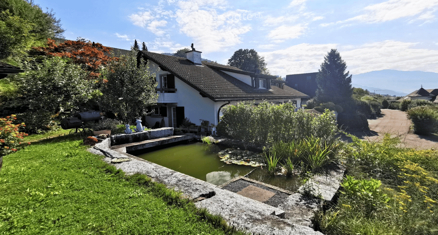 A serene garden area with a small pond, surrounded by lush greenery, a stone pathway, and a charming white house with a sloped roof in the background.