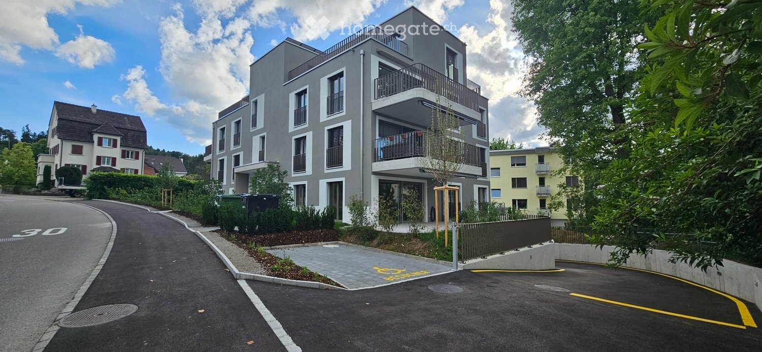 Modern multi-story apartment building with balconies, surrounded by greenery and a parking area.