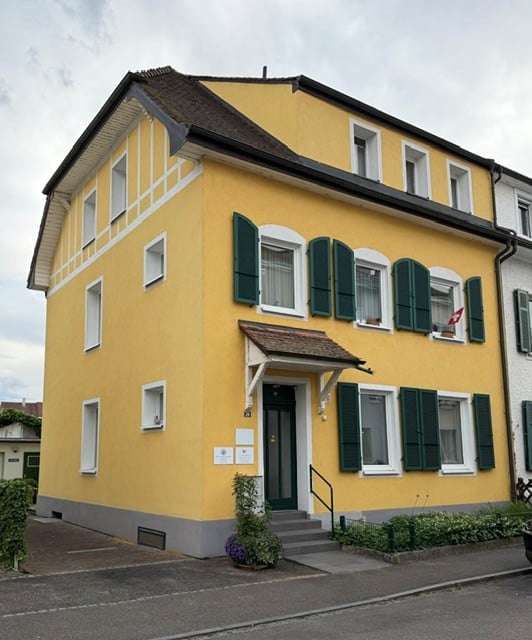 A two-story yellow house with white trim, green shutters, and a small porch with a staircase leading to the entrance.