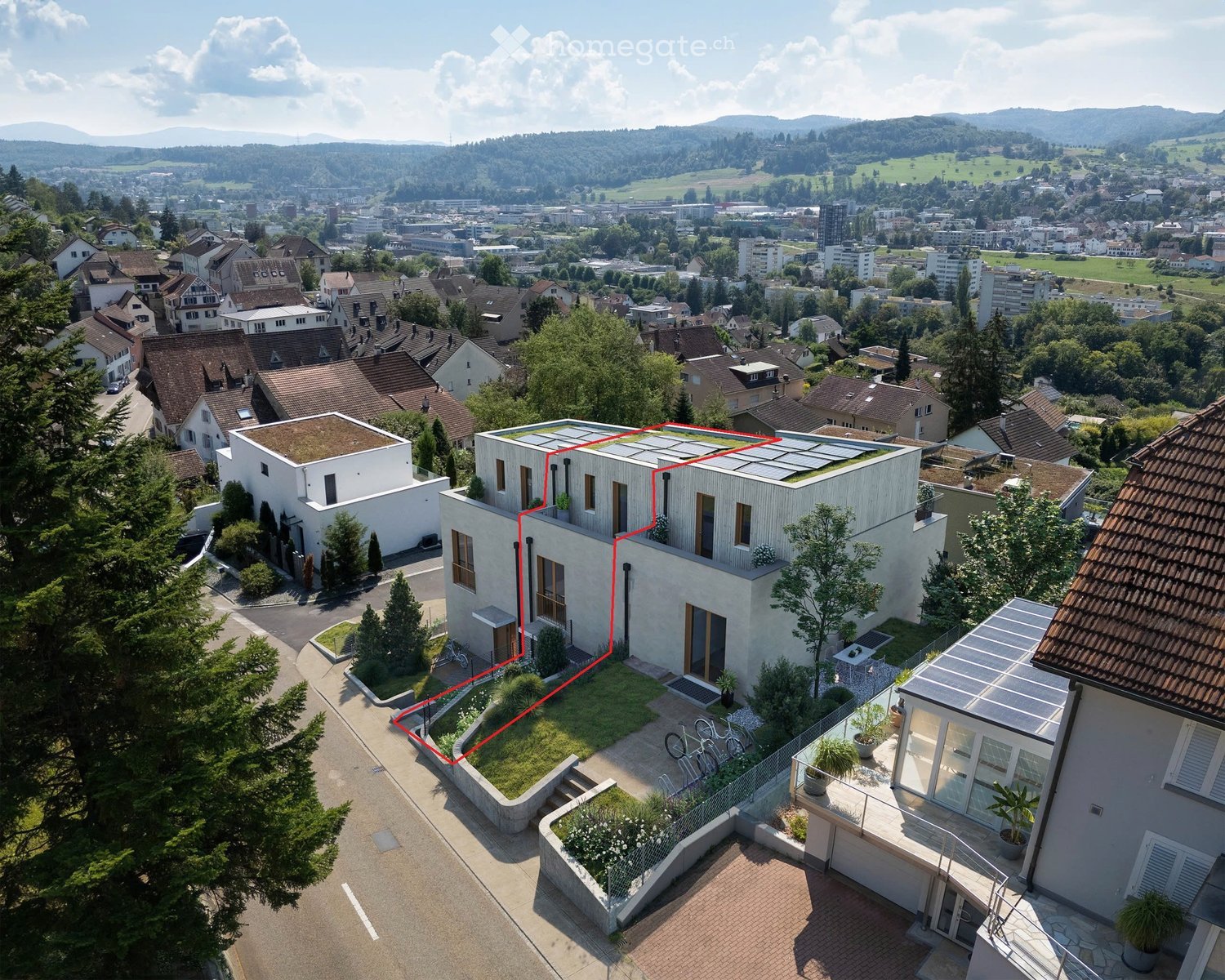 Aerial view of a modern, white house with a flat roof and solar panels, surrounded by greenery and neighboring houses in a suburban area with a scenic cityscape in the background.