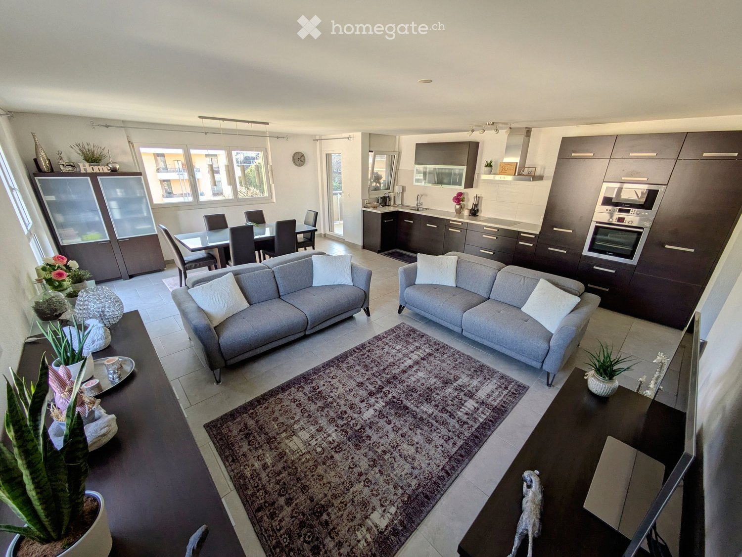 Open-plan living and dining area with grey sofas, coffee table, dining table, and modern kitchen with cabinets and appliances.