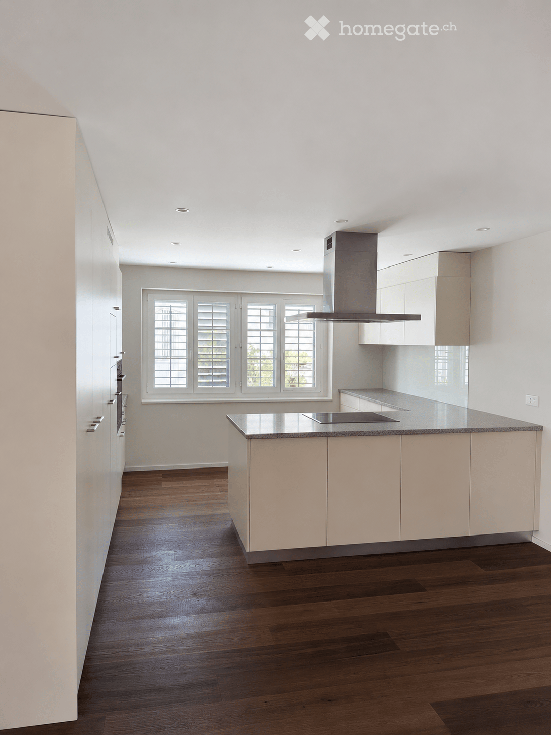 Modern kitchen with light-colored cabinets, a central island with a granite countertop, and a range hood above the stove. The floor is dark hardwood, and the room features large windows with shutters.