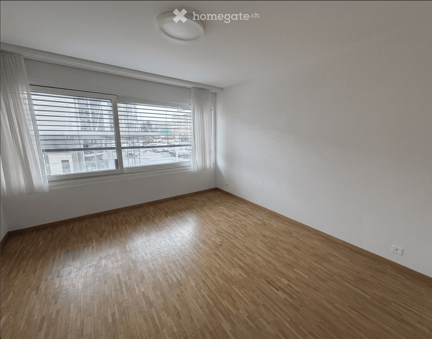 Empty room with wooden flooring, white walls, large window with blinds, and a ceiling light fixture.