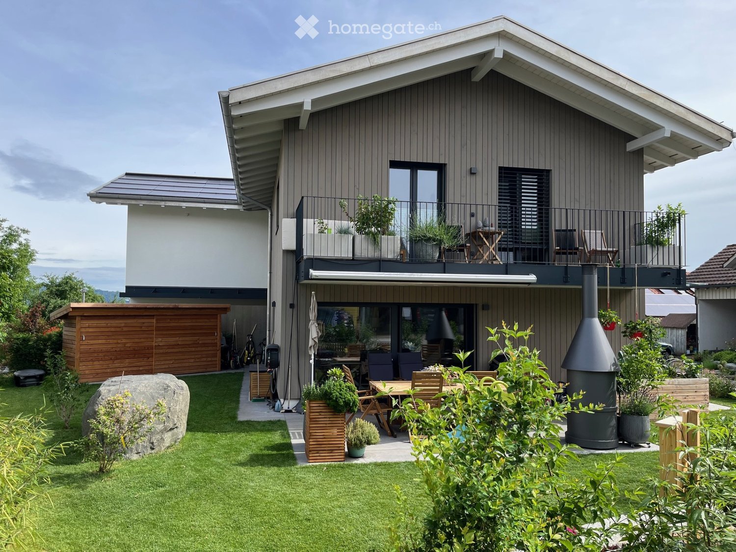 Modern two-story house with a well-maintained garden, featuring a patio area with outdoor furniture, a barbecue, and a balcony with additional seating.
