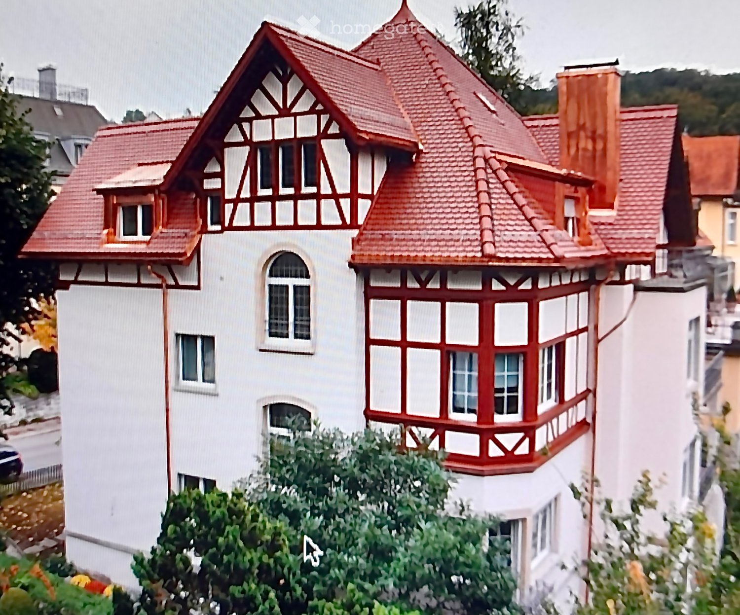 A two-story house with white walls, red wooden trim, and a red-tiled roof. The house features multiple windows, a chimney, and is surrounded by greenery.