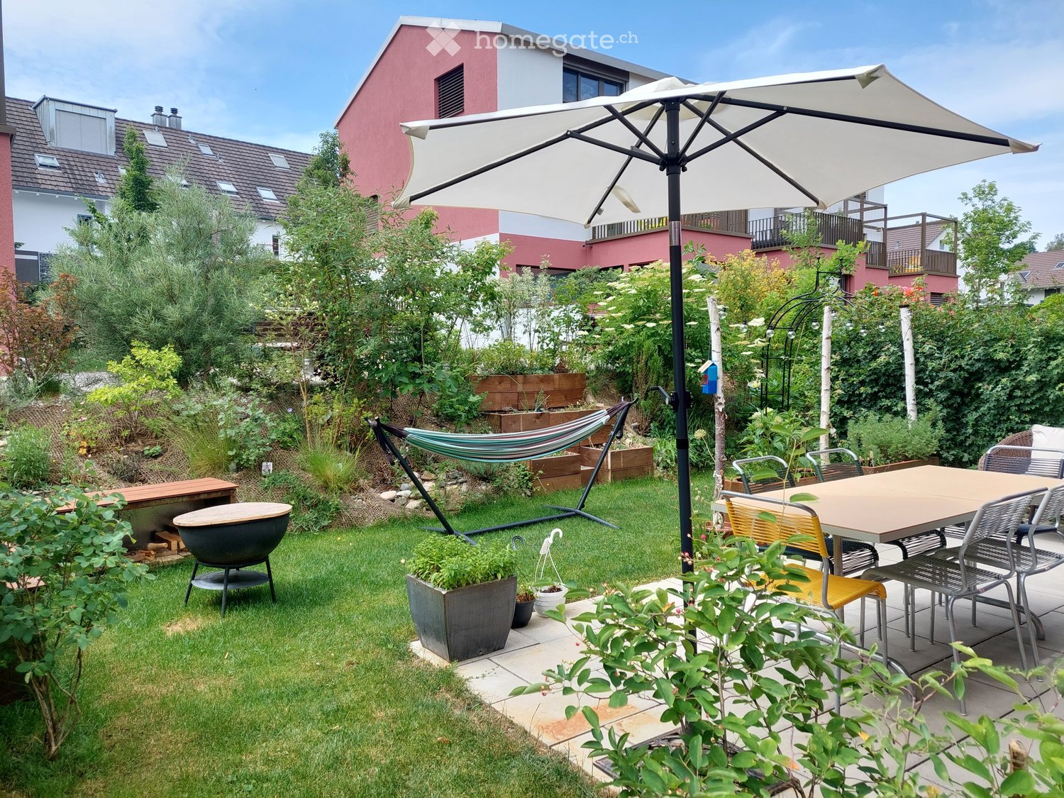 Well-maintained garden with a patio area, outdoor dining table, chairs, hammock, umbrella, and various plants and trees.