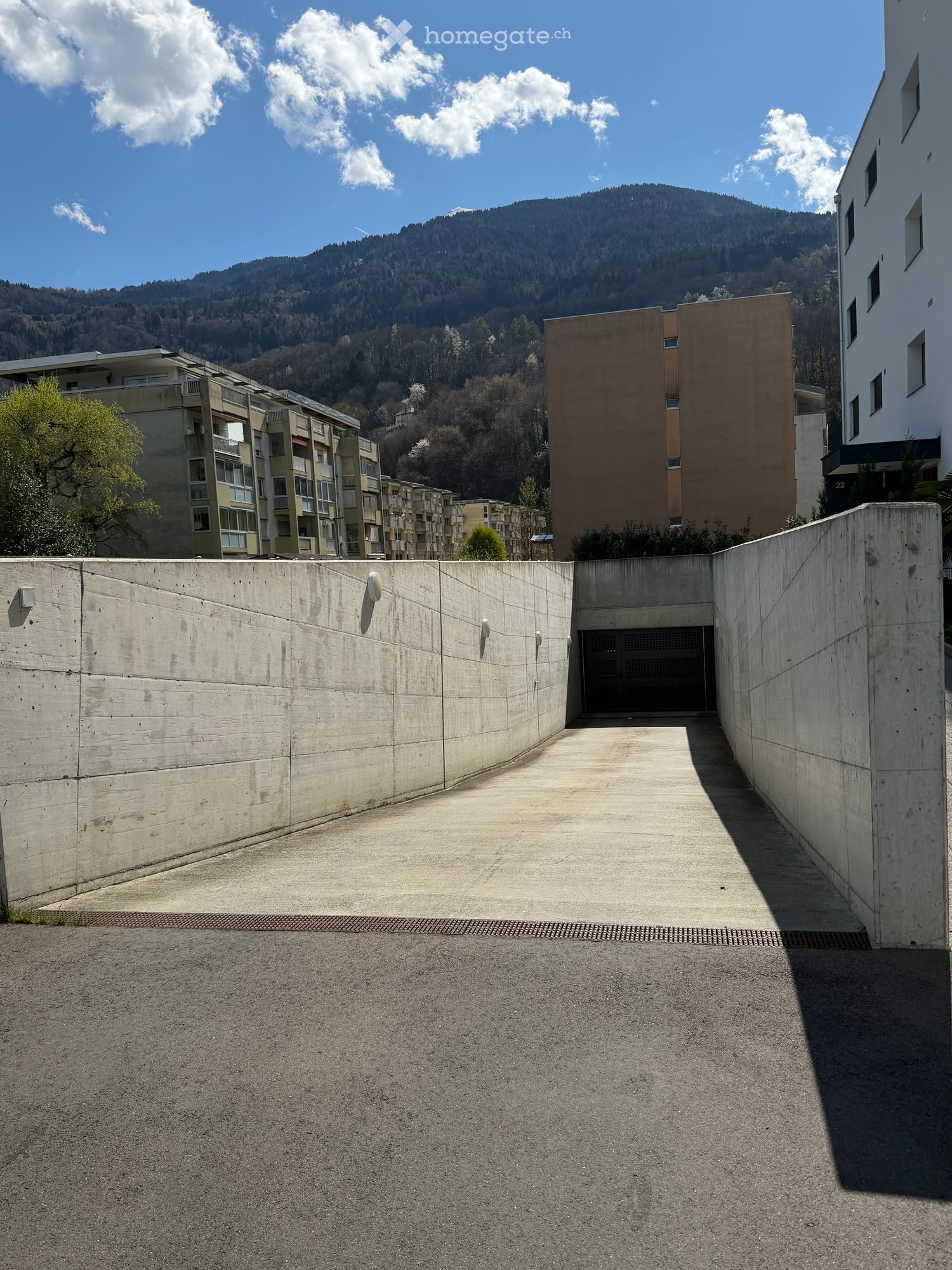 A modern residential complex with a raised ground floor entrance, surrounded by concrete walls and a scenic mountain backdrop.