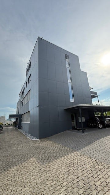 Building. Modern, multi-story building with a sleek, grey facade and large windows. Features a covered parking area with a few vehicles and a paved driveway.