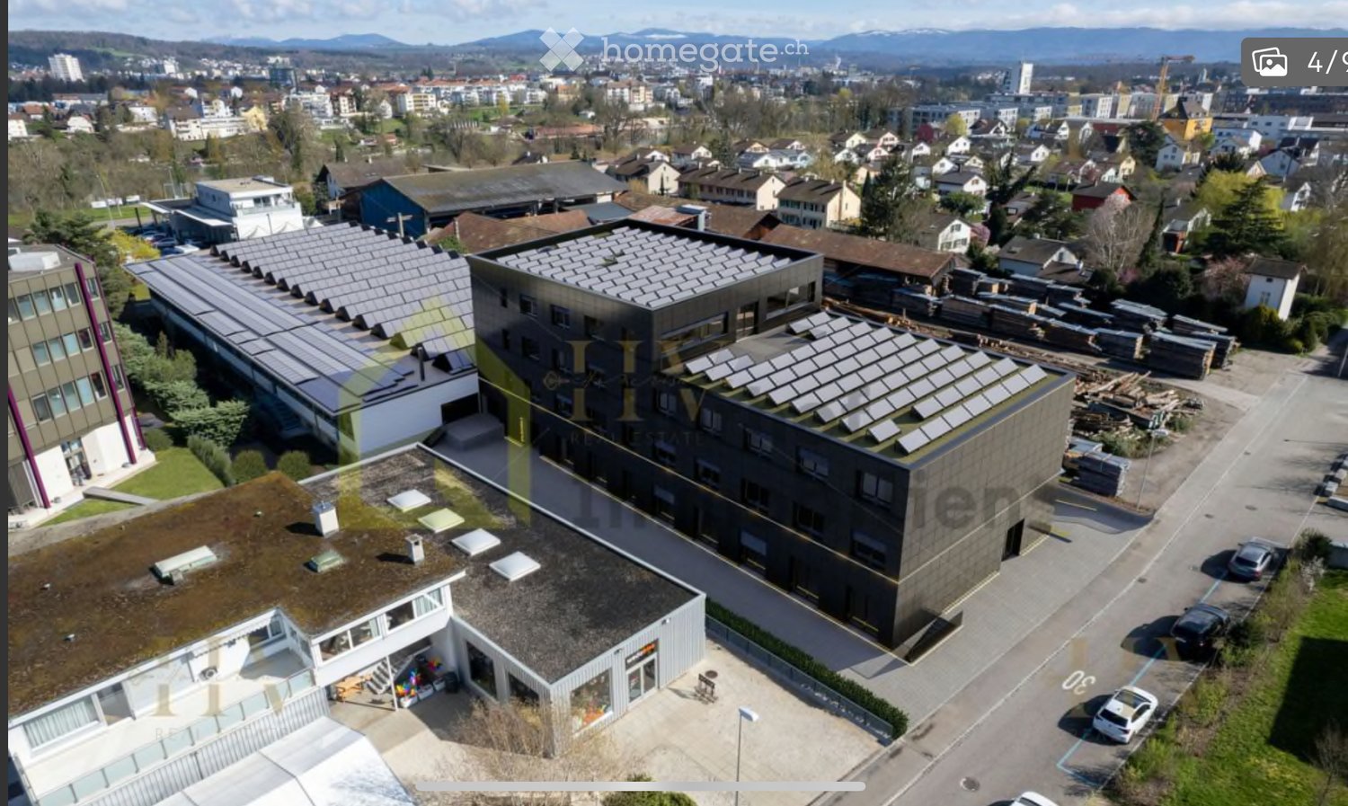 Aerial view of a modern building complex with solar panels on the roof, surrounded by other structures and a parking area.