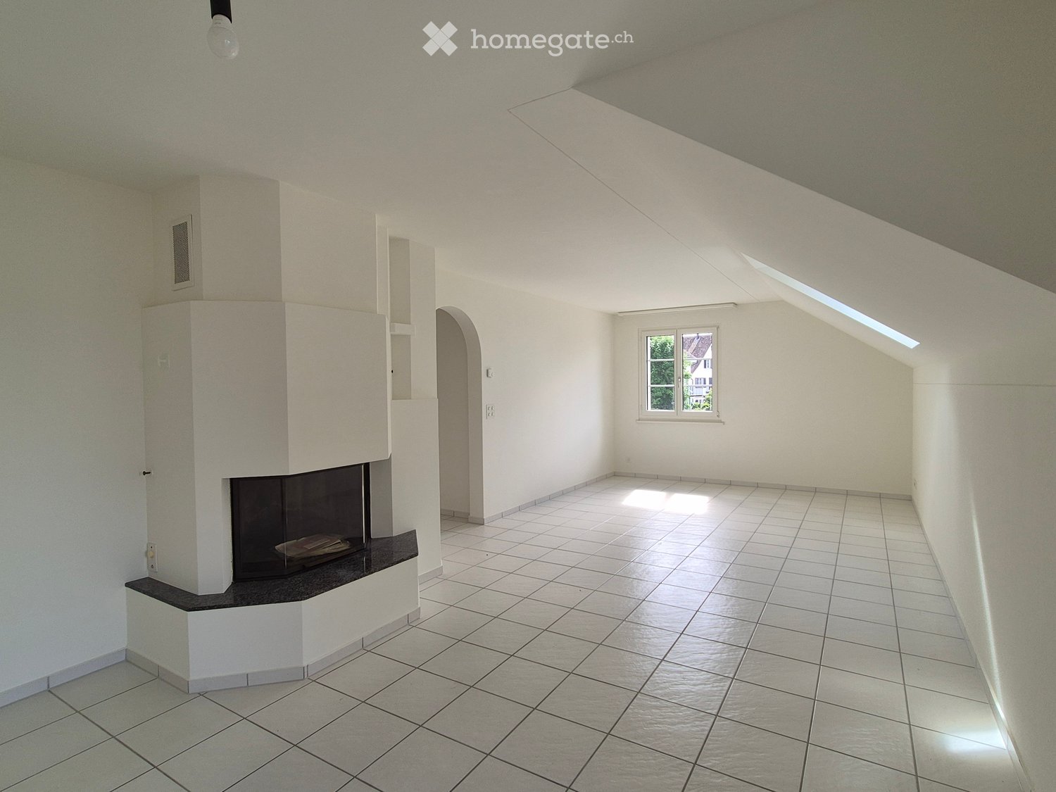 Empty room with white walls, tiled floor, and a built-in fireplace. There is a window on one wall and a skylight on the ceiling.