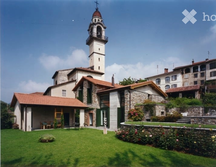 A historic church with a tall bell tower, surrounded by a well-maintained lawn and garden area. The building features stone walls, red-tiled roofs, and arched windows.