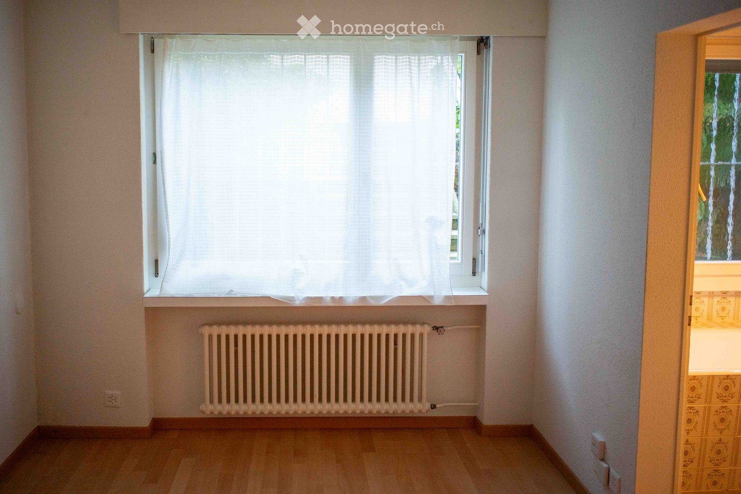 Empty room with wooden flooring, white walls, a window with white curtains, and a radiator below the window. The room is unfurnished with no visible furniture or personal items.