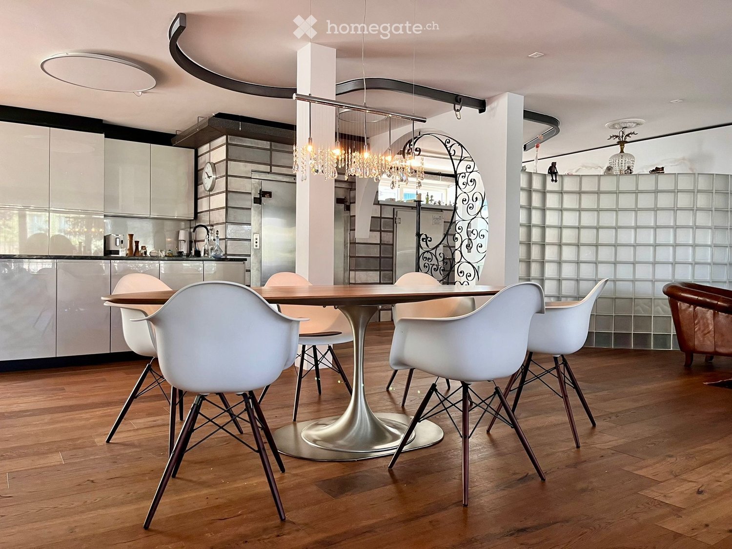 Modern dining area with a round wooden table, white chairs, and a sleek kitchen area in the background.