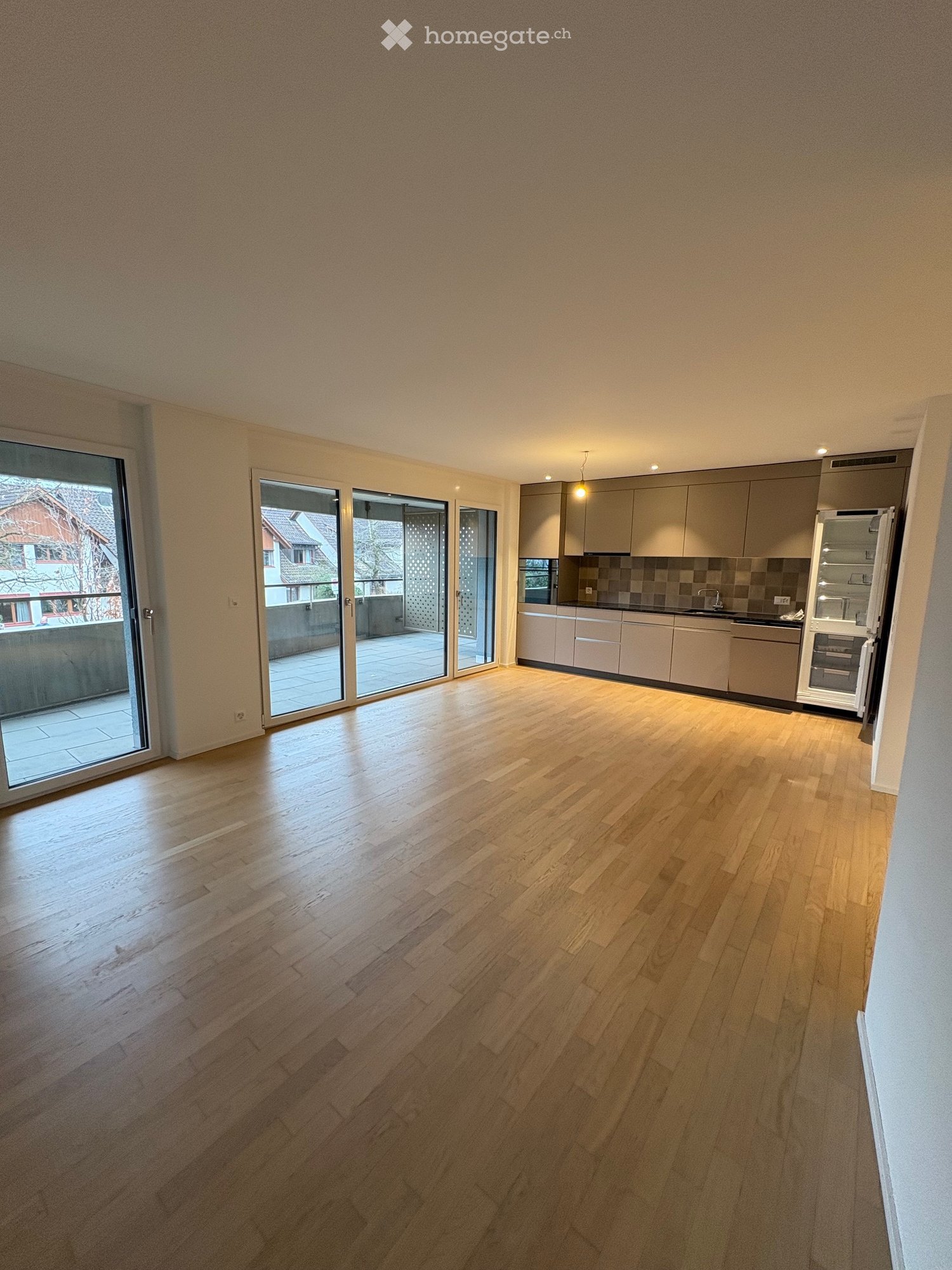 Empty room with wooden flooring, large sliding glass doors leading to a balcony, and a modern kitchen area with cabinets and appliances visible in the background.
