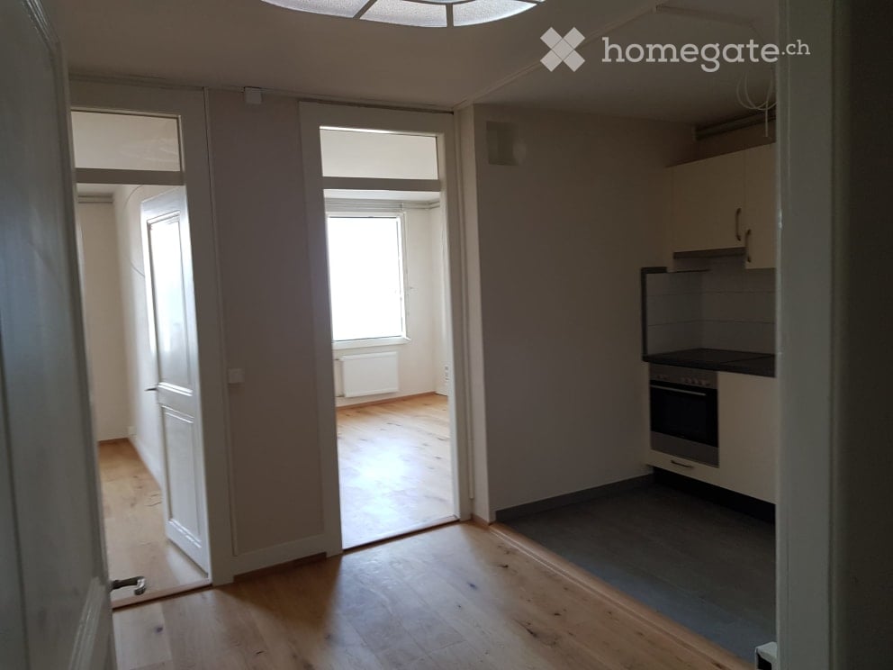 Empty room with wooden flooring, white walls, and a doorway leading to another room with windows. No furniture or equipment is present.