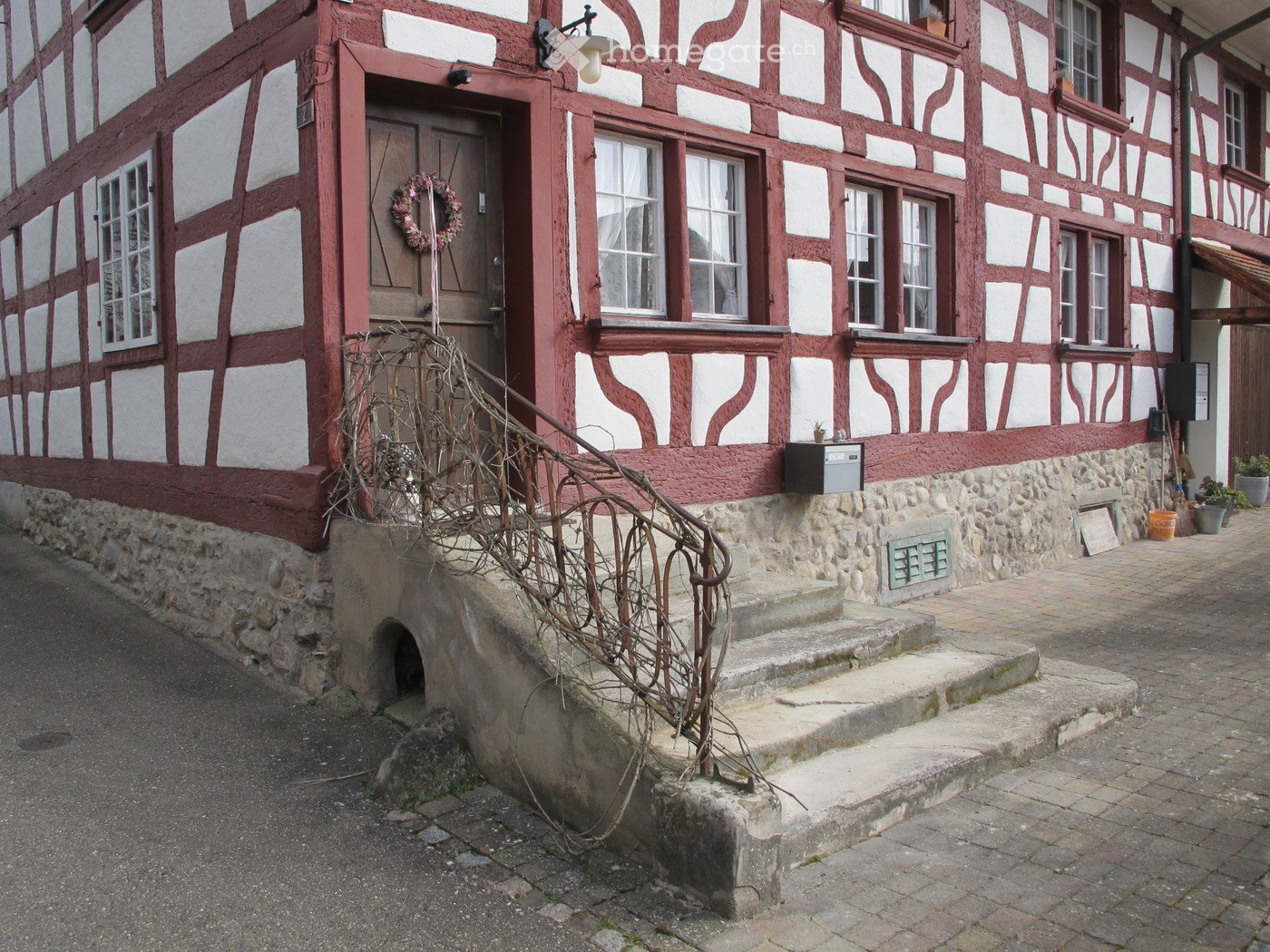 Traditional half-timbered house with red and white exterior, multiple windows, and a front door adorned with a wreath. A stone staircase leads up to the entrance, flanked by a metal railing and some potted plants.