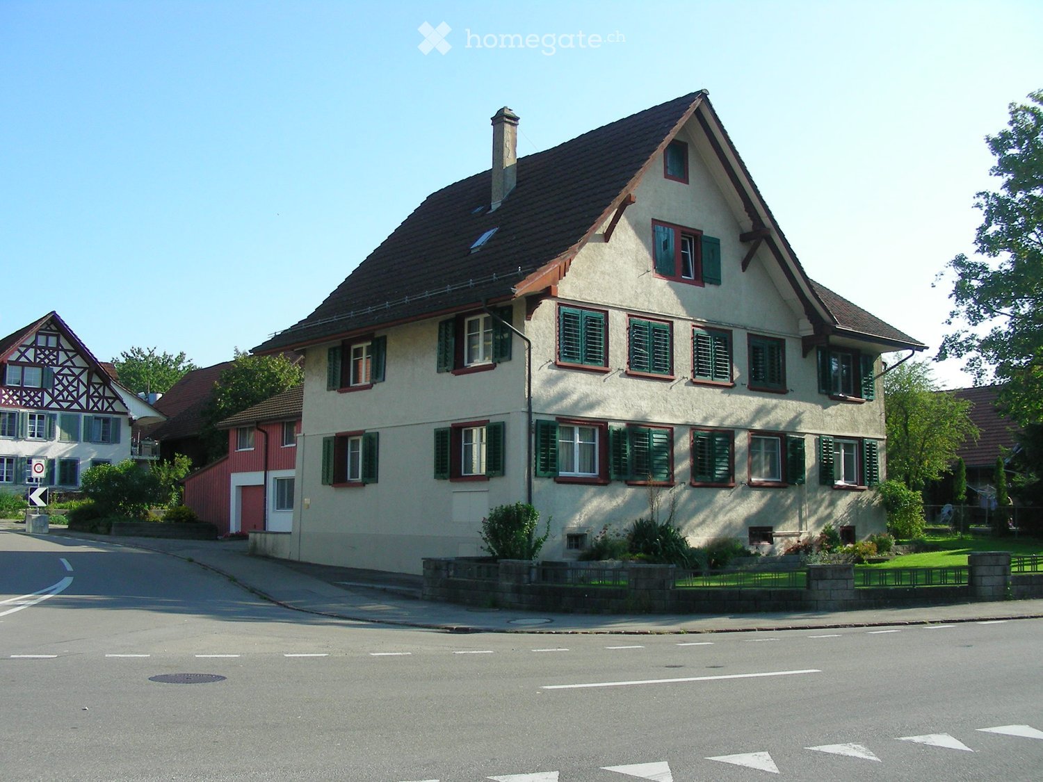 Two-story house with beige walls, green shutters, and a dark brown roof. The house has multiple windows and a chimney. There is a small garden area in front with a low fence.