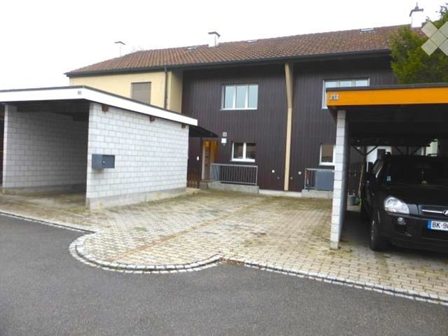 Two-story building with a garage and a covered parking area. The garage has a white brick facade, and the main building has a dark brown wooden exterior with white-framed windows.