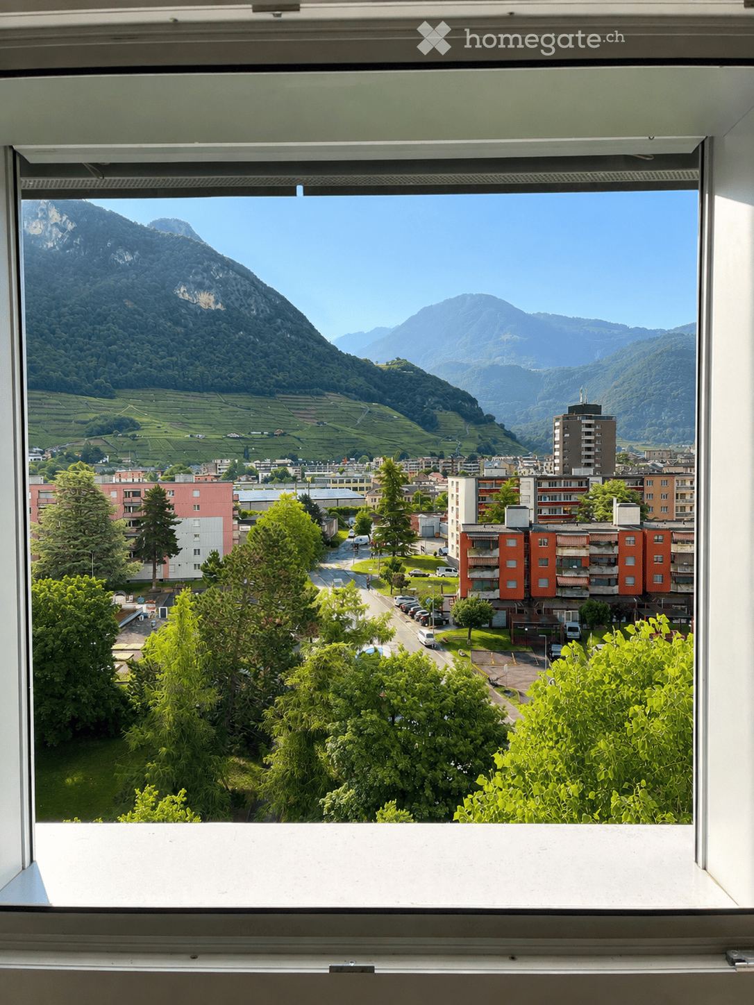 View from a window showing a town with buildings, trees, and distant mountains.