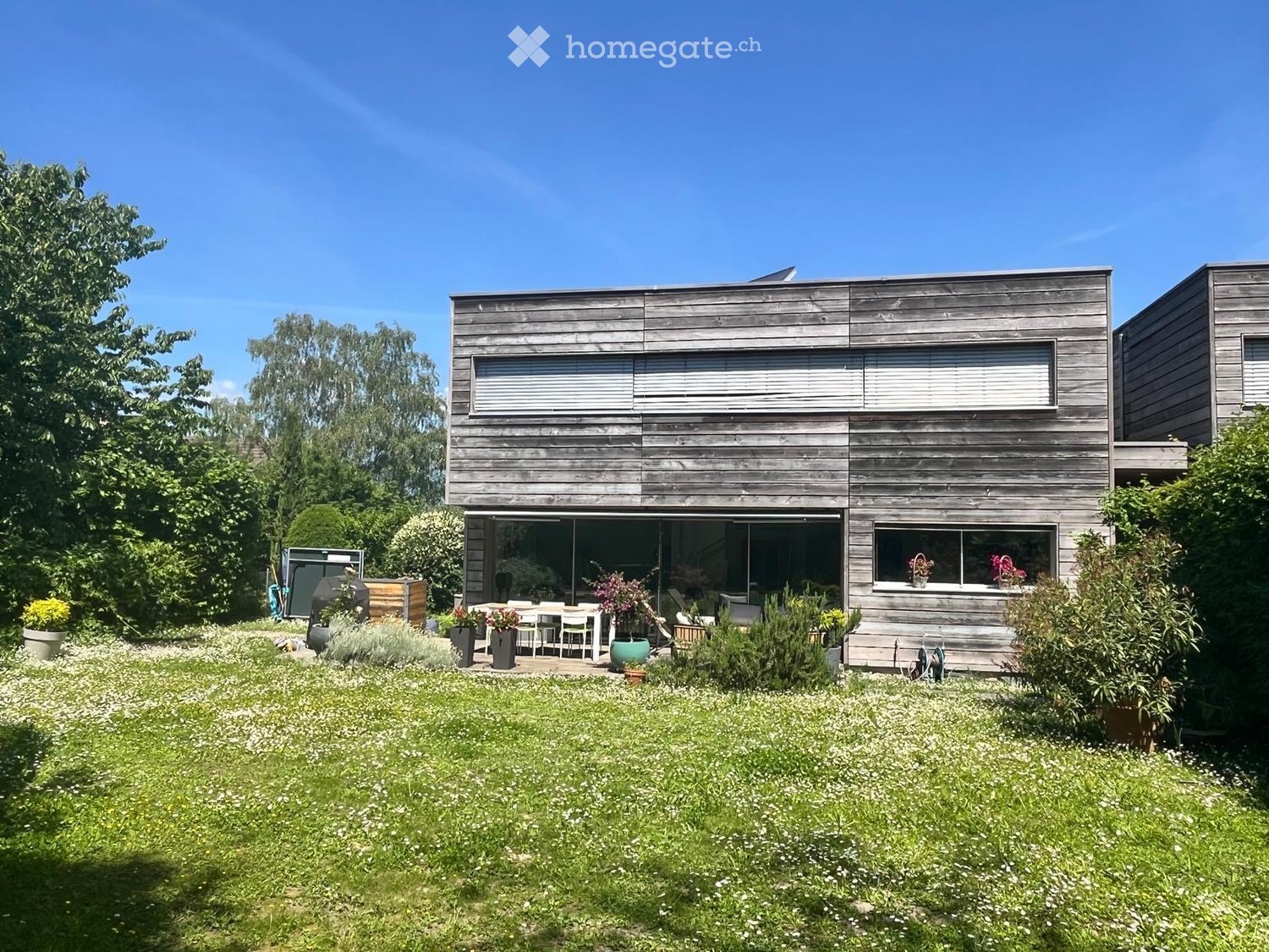 Modern two-story wooden house with large windows and a spacious garden area with a patio and outdoor furniture.