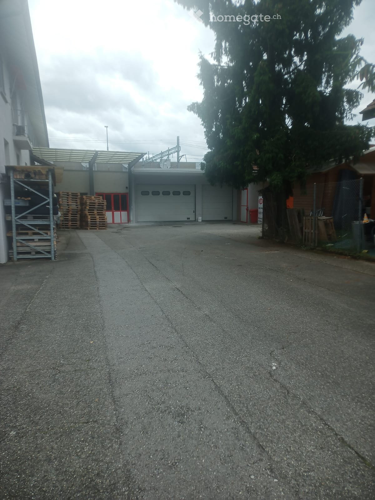 A paved driveway leading to a large industrial building with a garage and storage area, surrounded by trees and a fence.