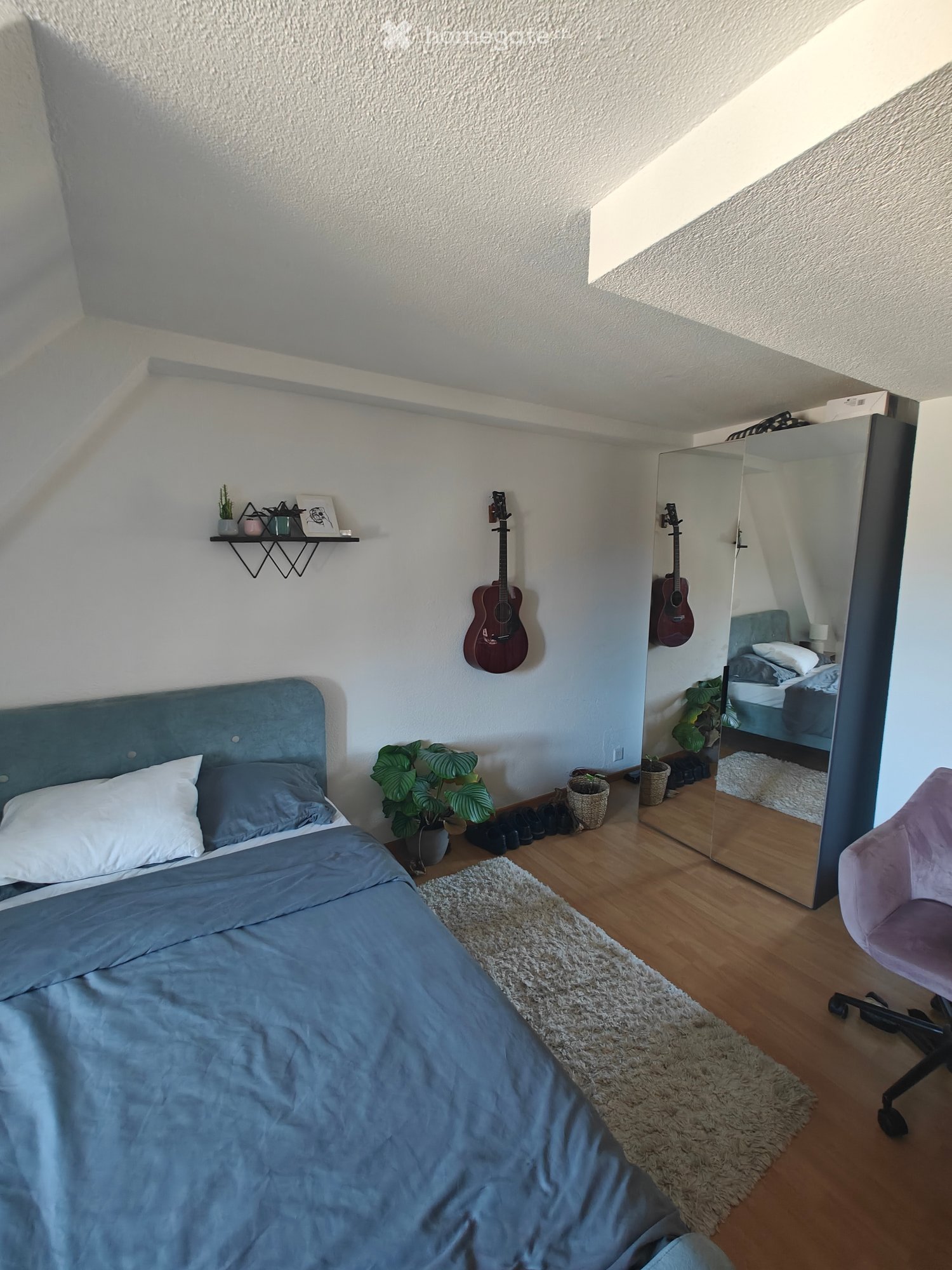 Cozy bedroom with a blue bed, wooden floor, large mirror, guitars, and a small shelf with decorative items.