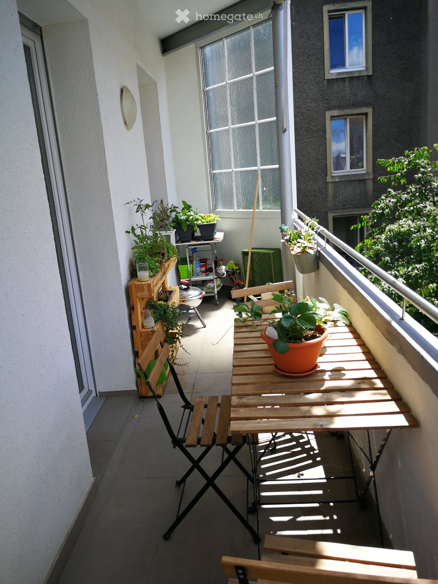 Balcony with wooden table, folding chair, potted plants, and railing overlooking greenery.