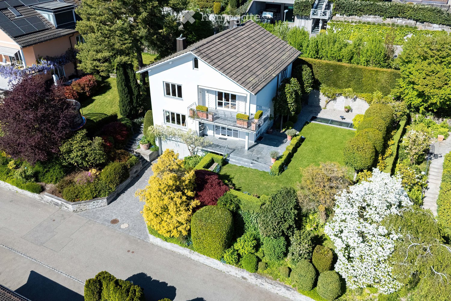 Aerial view of a two-story house with a well-maintained garden, featuring a driveway, a balcony, and surrounding greenery.