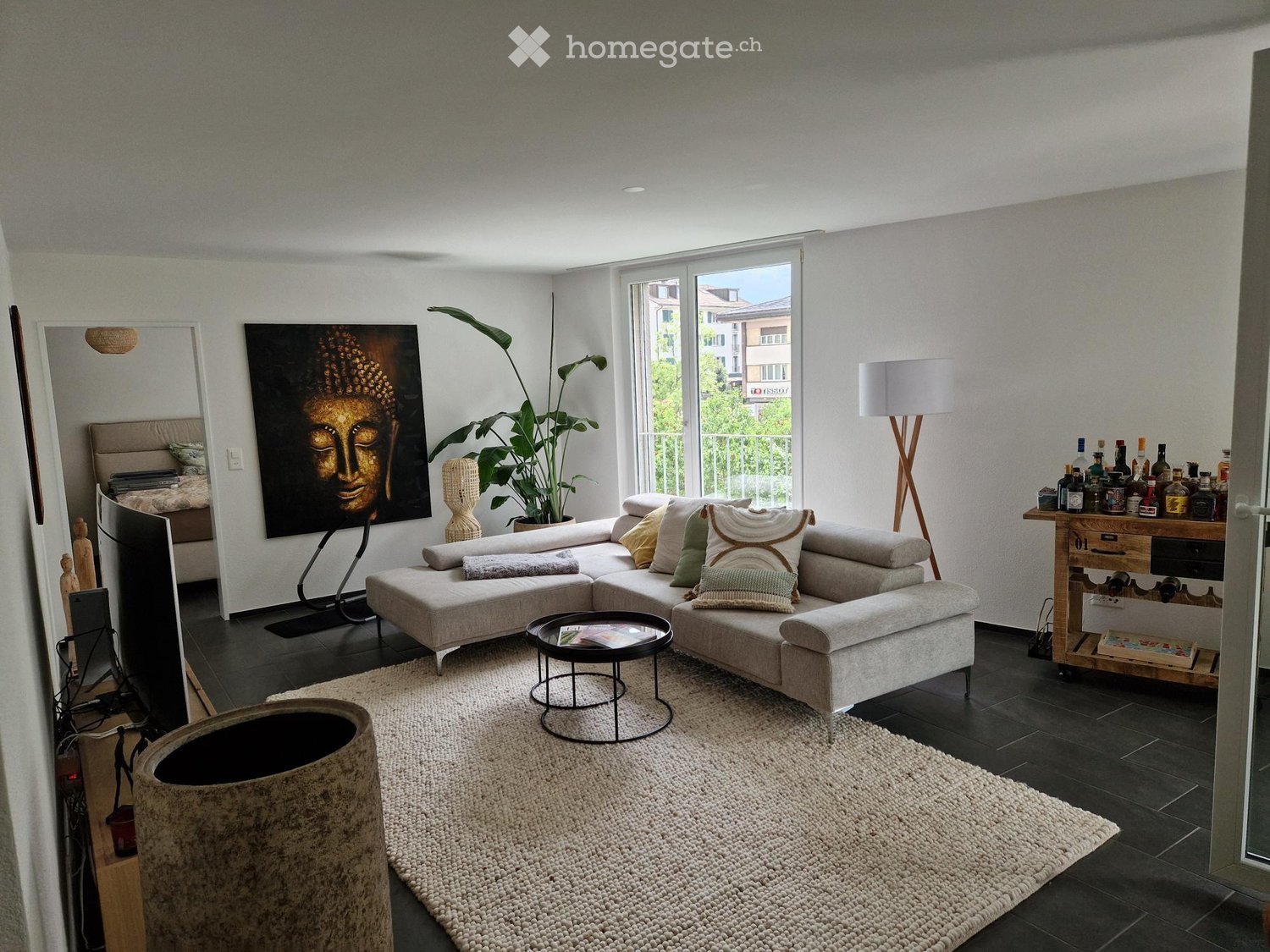 Modern living room with a grey sectional sofa, coffee table, floor lamp, large mirror, artwork, and a bar cart with bottles.