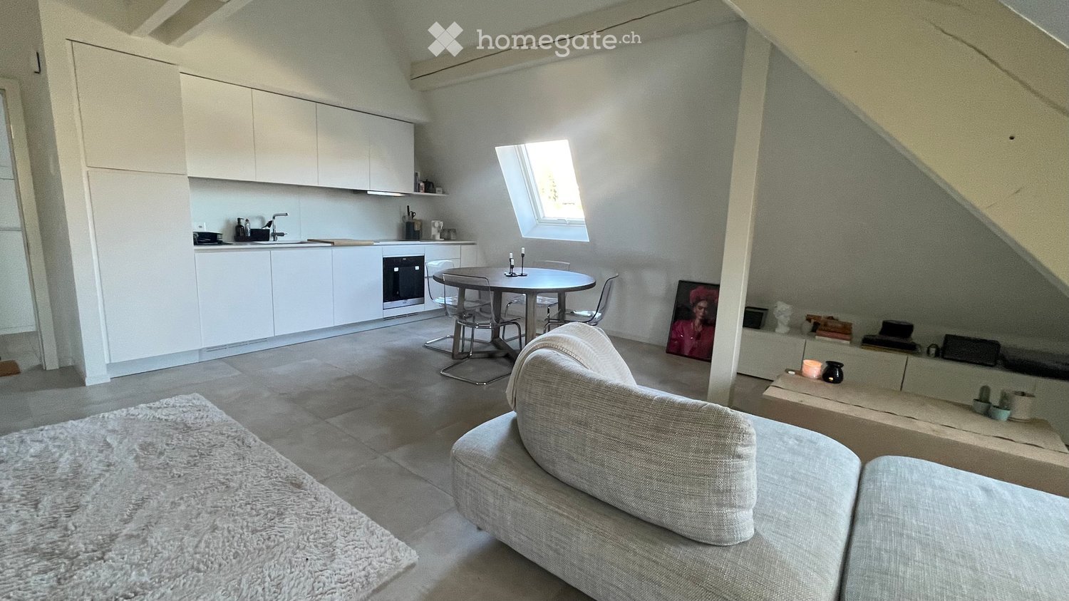 Open-plan living and dining area with a modern sofa, dining table with chairs, and a compact kitchen featuring white cabinets, a sink, and an oven. The space includes a large window, a fluffy rug, and decorative items such as a picture frame and candles.