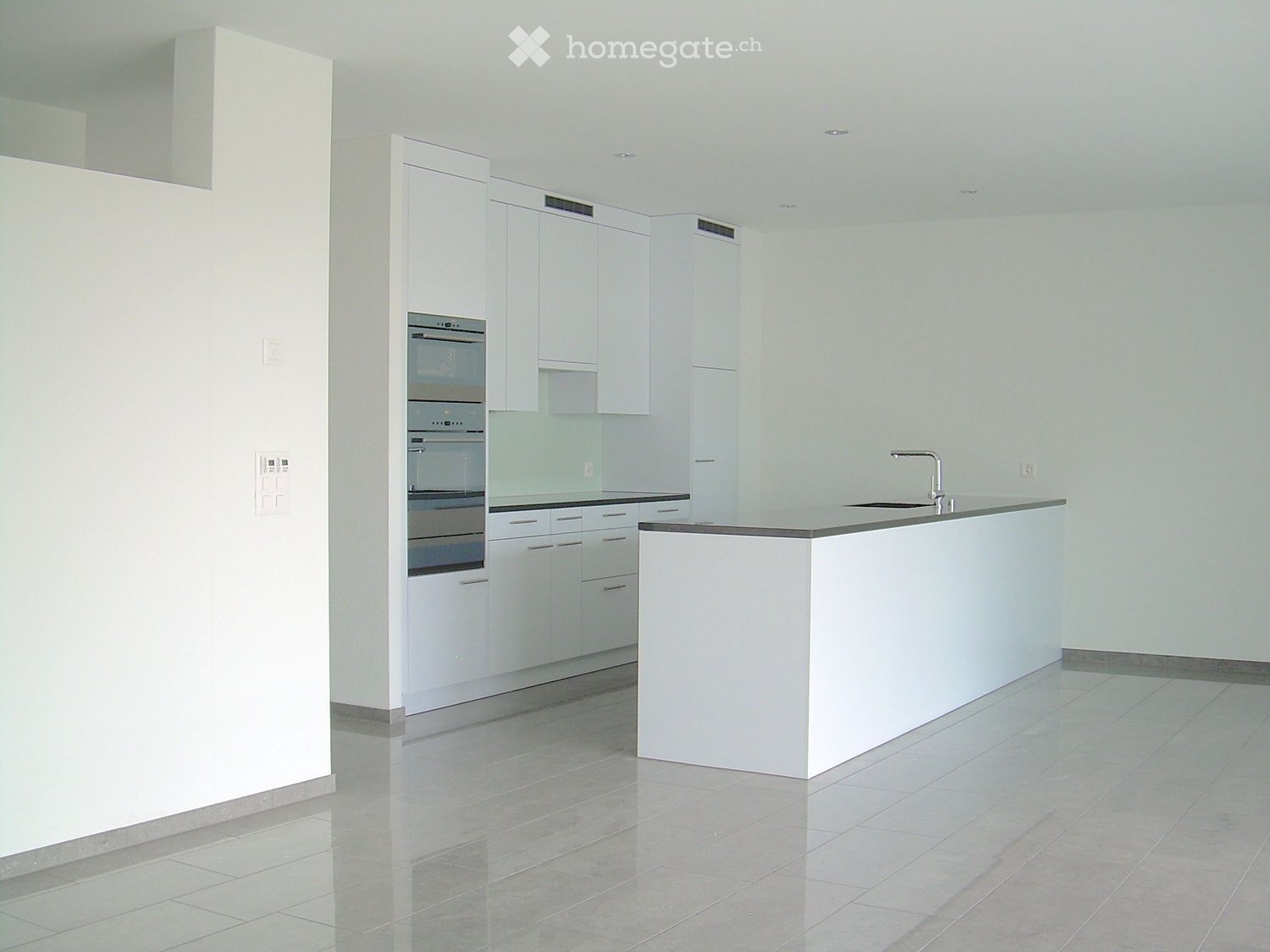Empty kitchen with white cabinets, a central island, and built-in appliances including an oven and microwave. The space features tiled flooring and neutral-colored walls.
