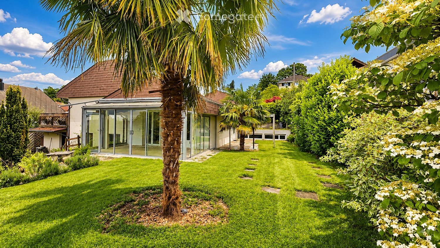 Well-maintained garden with a palm tree, neatly trimmed grass, and a modern house with large glass windows and a covered patio area.