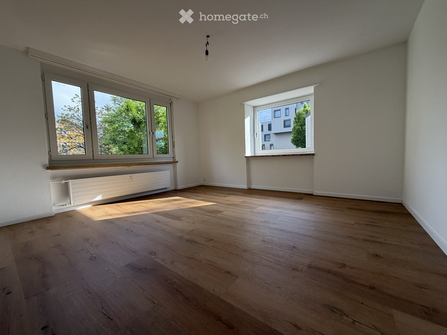 Empty room with wooden flooring, large windows, radiator, and natural light.