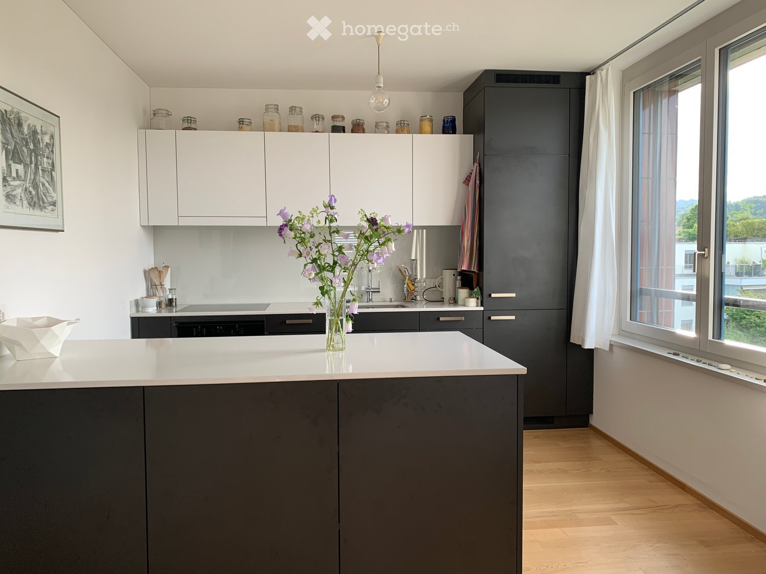 Modern kitchen with white and dark grey cabinets, a central island, and a vase of purple flowers on the counter. The kitchen features a sleek design with a hanging light fixture and large windows offering a view.